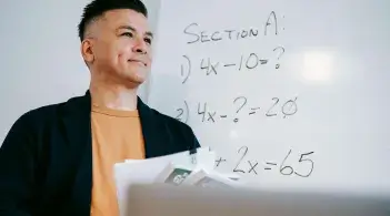 A Latino professor with short black hair, in a casual suit, smiles into the distance, in front of a whiteboard of equations