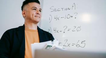 A Latino professor with short black hair, in a casual suit, smiles into the distance, in front of a whiteboard of equations