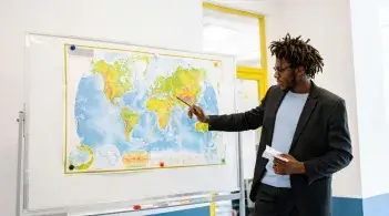 A Black man with short dreads and dark skin, dressed in a casual suit, points to a map tacked to a board in his classroom