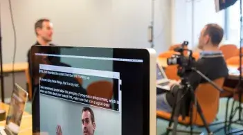 A flatscreen monitor shows live-captions of an interview taking place in the background