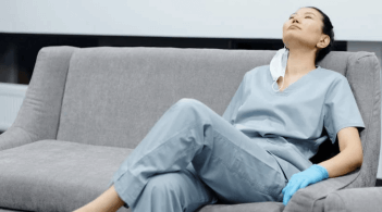 An Asian woman in scrubs and crocs, mask dangling from one ear, rests on a gray couch