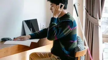 An almond-skinned college-aged man sits at a desk wearing headphones and running a hand over Braille text. A white cane stands nearby.