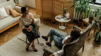 A copper-skinned woman with dark hair in a low bun and glasses, sits across from another woman, explaining her feelings