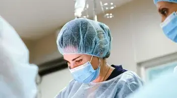 A white college-aged student in operating scrubs focuses intently on work on the operating table while a mentor looks on.