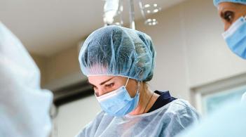 A white college-aged student in operating scrubs focuses intently on work on the operating table while a mentor looks on.