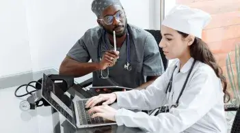 A Black doctor in scrubs sits at a desk watching a Latina medical student enter information on a laptop