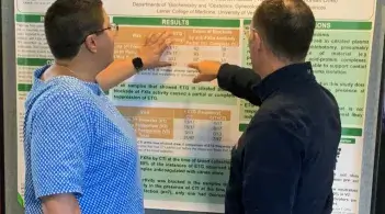 A person in a blue short sleeved shirt and a person in a black fleece jacket pointing at and discussing research data on a poster