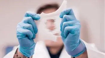 A researcher in a lab coat with blue gloves examines a stretchy material in the lab