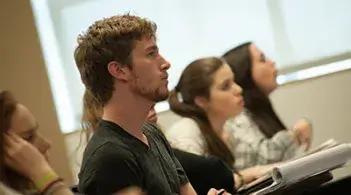 Students sit in a classroom listening attentively, with notebooks open in front of them.