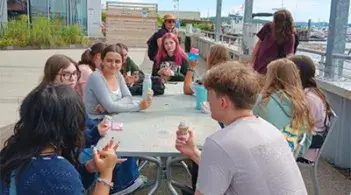 Students sit around an outdoor table eating ice cream and talking near a waterfront area.
