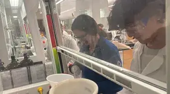 Two students wearing safety goggles work together at a chemistry lab bench behind a glass shield.