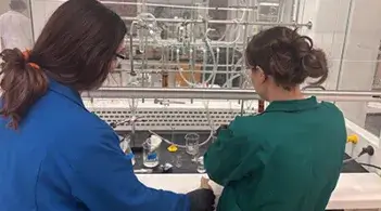 Two students in lab coats work side by side at a chemistry bench with glassware and equipment.