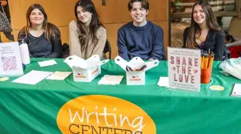 Four writing tutors sit behind a table, smiling, with handouts, Munchkins, and signs for Share the Love and a research survey. The Writing Centers orange oval wordmark is on the front of the green tablecloth.
