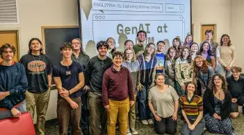 Interim UWC Director Holly Painter stands smiling in the middle of a group of Spring 2025 English 2795 students in front of a projector screen in a classroom. The projector screen reads, "GenAI at UVM."