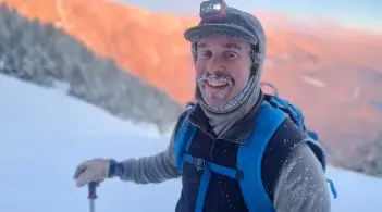 person with frost-covered facial hair and dressed in full winter gear stands on a mountainside in winter