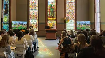 Audience attending an English department event in a chapel with stained glass windows.