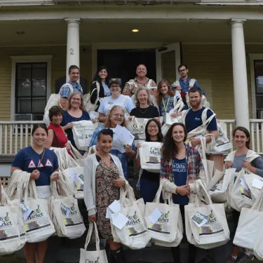 Group of volunteers holding reusable bags that read, "Welcome to the Neighborhood" stand on the porch steps of Pearl House.