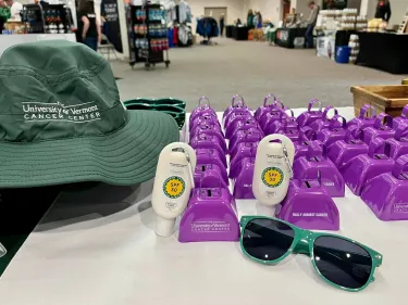 UVM Cancer Center branded sunscreen, pair of sunglasses, and some purple cowbells on a table