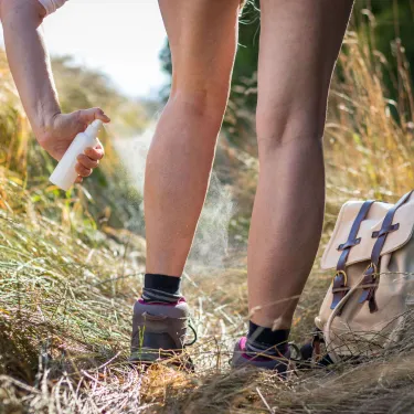Person standing in tall grass spraying bug spray on their legs.