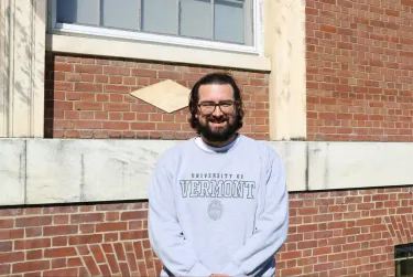 Person smiling in front of a brick building wearing a University of Vermont sweatshirt.