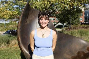 Person smiling outdoors in front of a large bronze sculpture on a sunny day.
