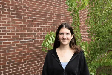 Person smiling in front of a brick wall with green leaves nearby.
