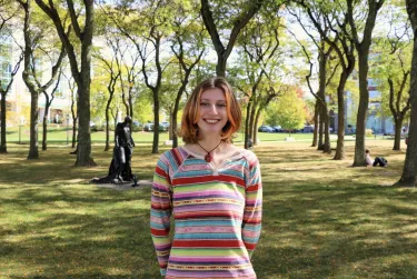 Person smiling outdoors in a park-like area with trees and a statue in the background.