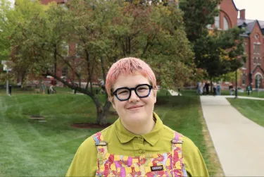 Person smiling outdoors on a campus pathway with trees and brick buildings in the background.
