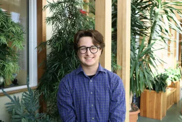 Person smiling indoors with plants and wooden beams in the background.