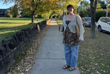 Person smiling on a tree-lined sidewalk holding books on a sunny day.