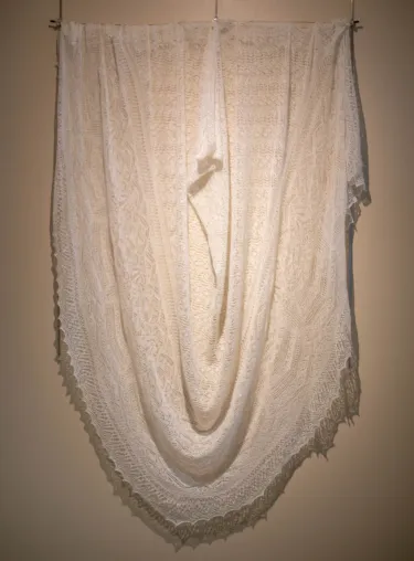 Hand-knit lace shawl with incredible ornamental detailing. The shawl is triangular in shape, with small designs made out of a very delicate, thin white yarn. The shawl is draped and hung across the wall under an overhead light to accentuate the texture. 