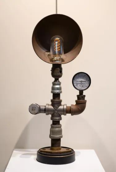 Functional lamp made of repurposed materials, including a circular copper piece, piping and a pressure gage. The base of the lamp is a vertical pipe which has a pressure gage attached on its right. The light bulb screws in at the top of the pipe with a copper circle behind it, which is illuminated when the lamp is plugged in. 