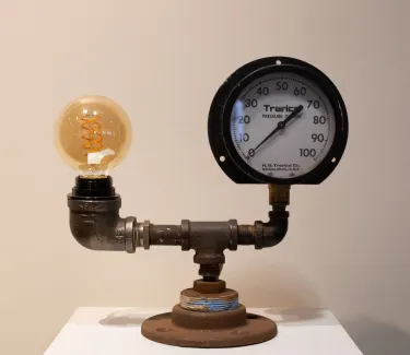  Functional lamp made of repurposed materials, including pipe and a pressure gage. The pipe on this lamp goes horizontally, with a pressure gage on the right and the lightbulb on the left. There is no shade. 