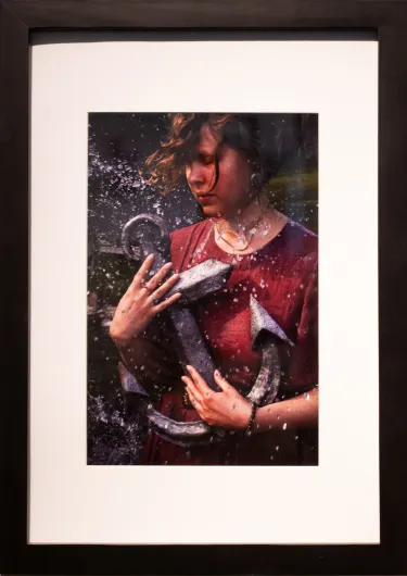  Digital photograph, archival print. Matted and Framed 15.5 X 12.5in. A woman dressed in red with closed eyes and brown hair blowing across her face holds a large, grey metal anchor as water droplets splash across her figure. 