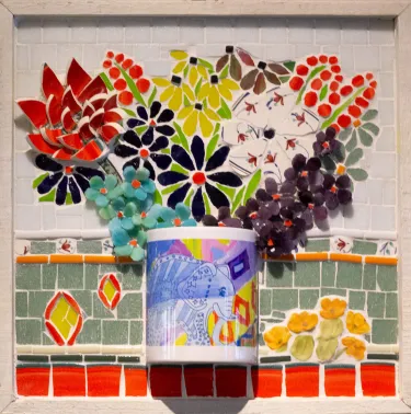 3-dimensional mosaic art panel in the Pique Assiette style depicting a bouquet of flowers in a mug vase. The bouquet is centered in mosaic, with a variety of flowers. On the left, there is a red flower made with large tear-shaped tiles, to it's right there are large black flowers with red centers, above those are black-eyed susans made with yellow tiles, to the right of those there's a white flower made from floral patterned tile. The top half of the background is white tile and the lower half is grey green