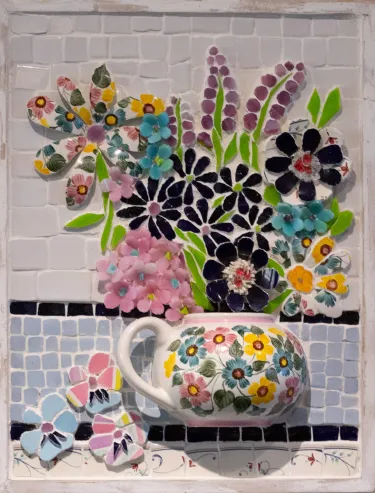3-dimensional mosaic art panel depicting a bouquet of flowers in a floarl teapot vase. A round handled vase with painted floral designs holds flowers made of tile, with pink hydrangea-like flowers on the bottom left, a collection of balck daisies in the center, a flower to the left with the same floral pattern as the vase, lavender in the top right, and another flower made of the floral china on the right. The background is white with a strip of blue tile on the bottom half. The vase rests on a row of decor