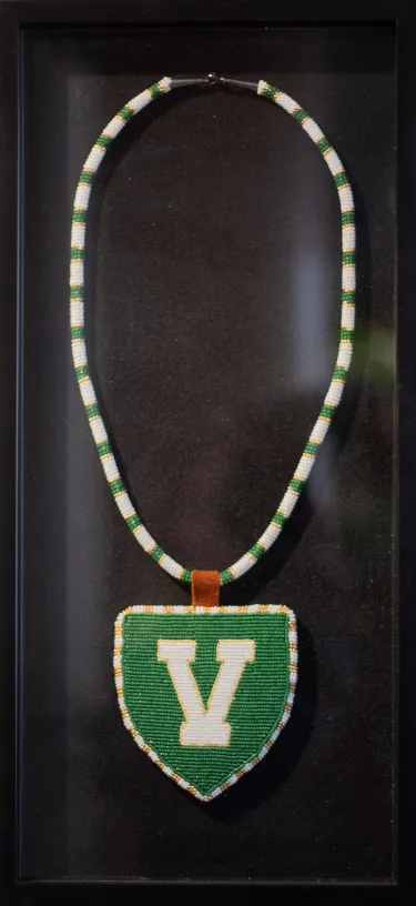 Beaded medallion with UVM's "V shield" logo in green and white. White and gold beads border the triangular green beaded logo to make the pendant. A leather strap connects the pendant to a chain made of white, gold and green beads which alternate so that six rows of white beads are followed by one row of gold, three rows of green and another row of gold, then the pattern repeats.