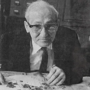 an elderly Seymour with glasses in a suit sits at a desk, examining plant specimens