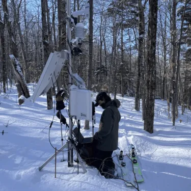 a person in winter gear examining a weather monitoring station in the snowy woods