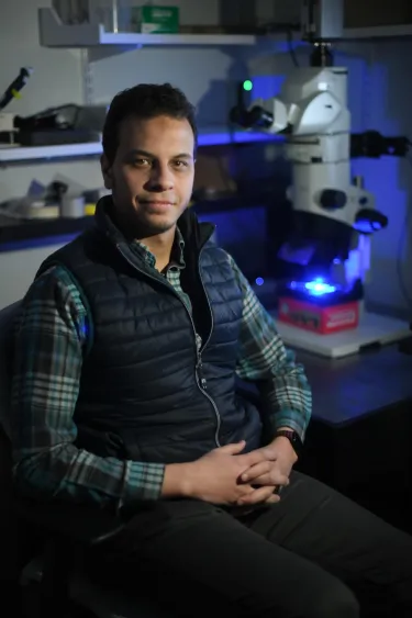 man wearing a plaid shirt and puffy vest sitting in a dark lab with hands folded in lap