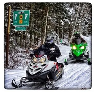Photograph of two people riding snowmobiles on a snowy forest trail, with one snowmobile in the foreground and another following behind. A green trail sign with directions and distances is visible on the left side, indicating a marked snowmobile route through a wooded area.