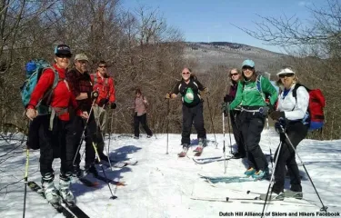 Photograph of eight people standing on a snowy trail with skis and poles, dressed in winter hiking gear and backpacks. Background shows leafless trees and distant hills under a clear sky, indicating a group ski outing in a natural, mountainous area of Vermont.