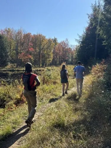 Three college students walking along a trail that runs through a field with trees on a sunny day.