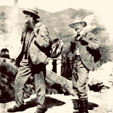 Two men in weathered suits, boots, and broad-brimmed hats, Pringle with beard and carrying plant specimens in case, Lozano on right with mustache and backpack