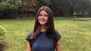 Portrait of a student standing outdoors in a grassy yard surrounded by trees and greenery.