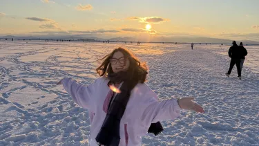 Portrait of a student standing on a snow-covered landscape at sunset, with mountains in the distance and other people in the background.