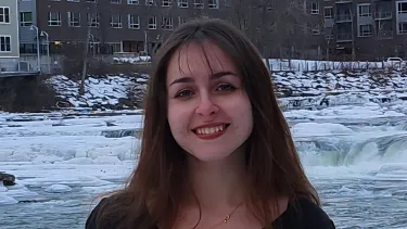 Portrait of a smiling student standing in front of a partially frozen river, with snow-covered rocks and buildings in the background.