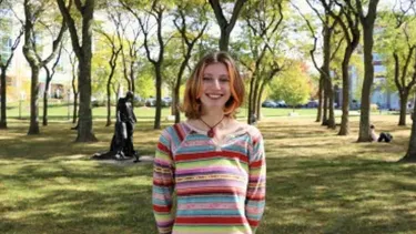 Student standing in a tree-lined park with grass and campus buildings in the background.