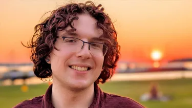 Portrait of a smiling student with curly hair and glasses outdoors at sunset, with a grassy field and waterfront in the background.
