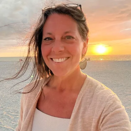 A white woman with long brown hair, pulled back but with some tendrils loose, stands on a white sandy beach with the sun setting behind her. She has glasses pushed up on the top of her head, she is smiling.